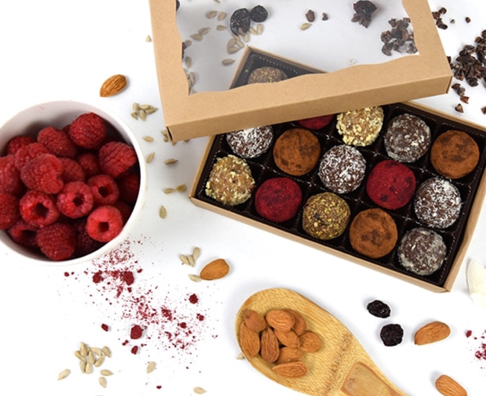 An open kraft gift box of fifteen bliss balls beside raspberries and almonds